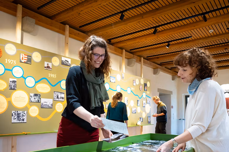 Ausstellungsraum im Schloss Lenzburg: Zwei Frauen stehen an einem Tisch und stöbern in alten Fotos, im Hintergrund weitere Ausstellungselemente.