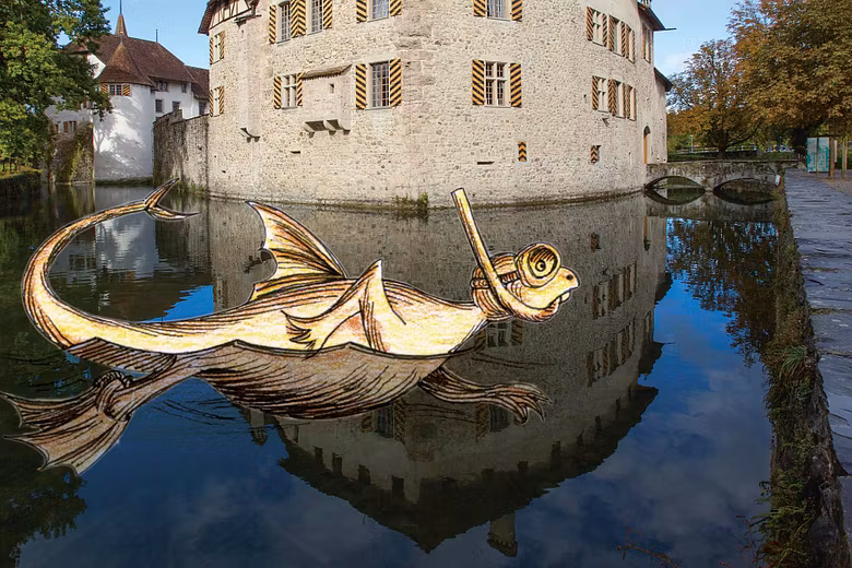 Aussenaufnahme von Schloss Hallwyl mit Aabach im Vordergrund. Im Wasser schwimmt ein Drachentier mit Schnorchel (Illustration), am Himmel ist ein Luftschiff zu sehen (Illustration)
