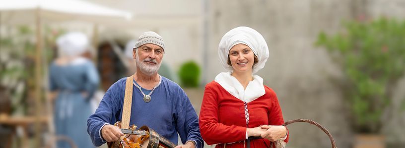 Eine mittelalterlich gekleidete Hofdame mit einem Musiker (mit Drehleier in der Hand). Beide blicken direkt in die Kamera.