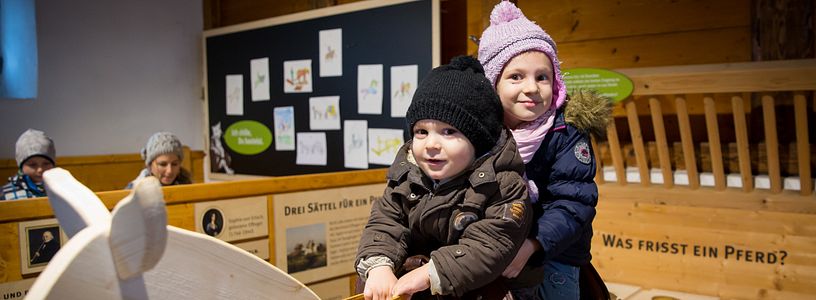 Mädchen und Junge reiten auf einem Holzpferd im Pferdestall auf Schloss Wildegg