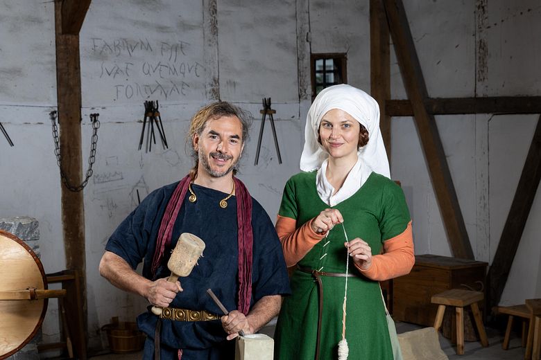 Ein römischer Steinmetz und eine mittelalterlich gekleidete Frau mit Wolle. Beide stehen in einem Werkraum und blicken direkt in die Kamera.