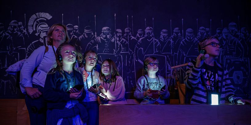 Eine Familie in einem dunklen Raum; die Displays der Audiogeräte um den Hals leuchten mystisch im Dunkeln. An der Rückwand Silhouetten von Legionären.