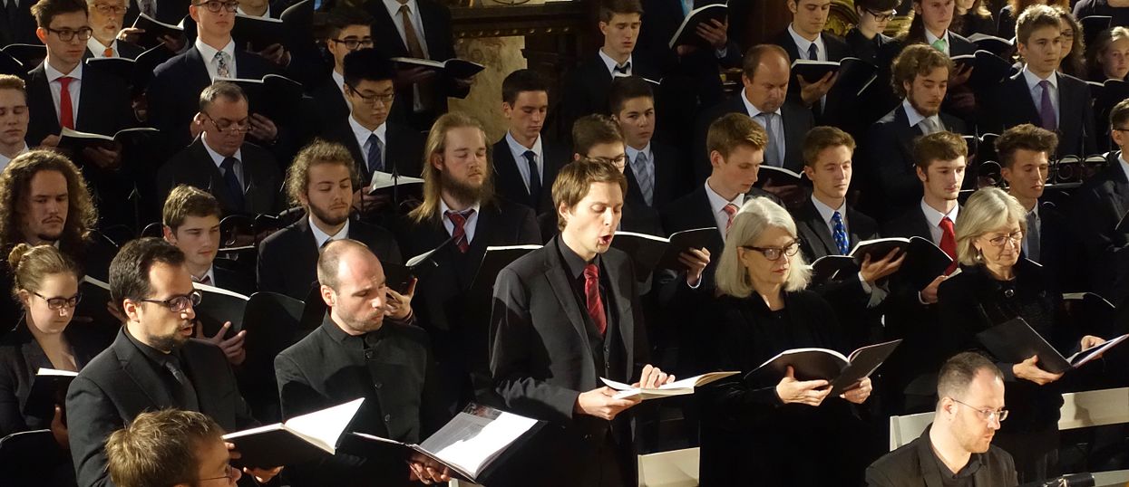 Viele Menschen, alle chic gekleidet und mit einem Notenbuch in der Hand am singen. Sie sind in mehrere Reihen aufgestellt. 