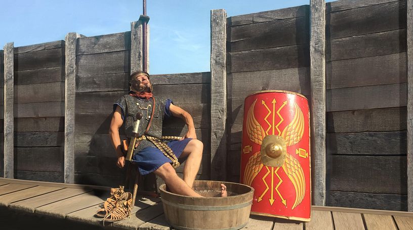 Ein römischer Legionär sitzt auf einem Stuhl und blickt entspannt in die Sonne. Seine Füsse liegen in einem Bottich mit Wasser.