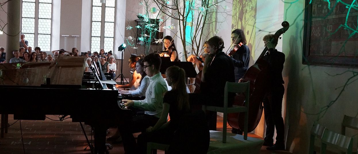 In der rechten Hälfte des Bildes stehen Schülerinnen und Schüler und spielen verschiedene Instrumente. In der linken Hälfte im Hintergrund sitzt das Publikum.