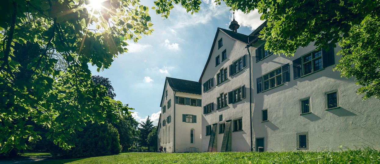 Aussenaufnahme Kloster Wettingen mit blauen Himmel, Sonne im Gegenlicht und einer grünen Wiese im Vordergrund.