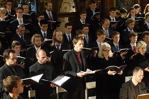 Viele Menschen, alle chic gekleidet und mit einem Notenbuch in der Hand am singen. Sie sind in mehrere Reihen aufgestellt. 
