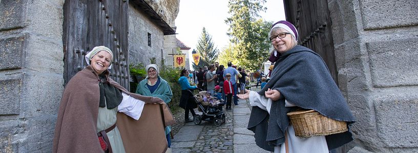 Zwei historisch gekleidete Freuen stehen vor dem Tor von Schloss Lenzburg und bitten Besucher mit einer einladenden Geste herein.