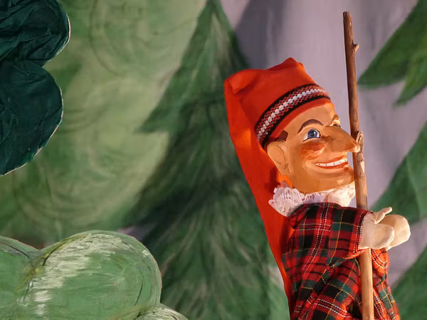 Kasperlefigur in roter Tracht mit einem Stock, die in einer Wald-Szenerie des Puppentheaters steht.