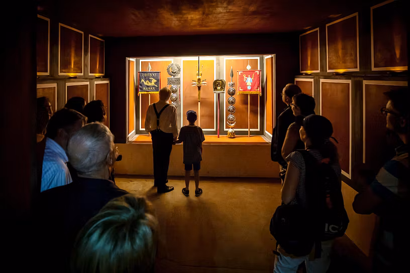 Mehrere Personen stehen im Ausstellungsraum "Fahnenheiligtum" im Legionärspfad Vindonissa und blicken in eine hell erleuchtete Vitrine mit Fahnen.