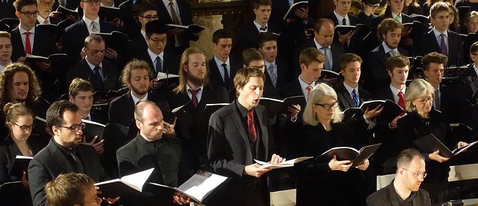 Viele Menschen, alle chic gekleidet und mit einem Notenbuch in der Hand am singen. Sie sind in mehrere Reihen aufgestellt. 