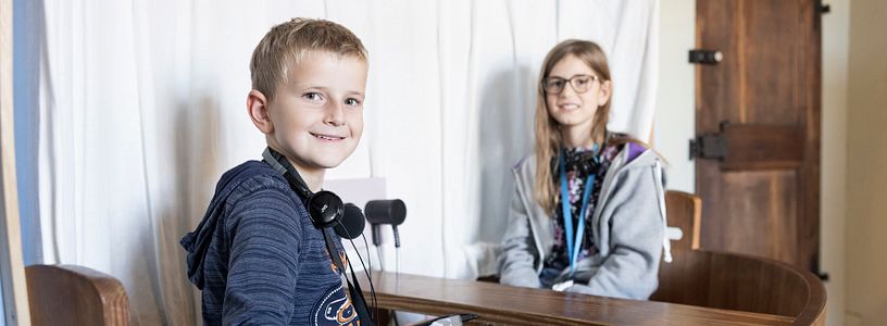 Ein Bub und ein Mädchen sitzen mit Kopfhörern in einem kleinen Schiff in der Ausstellung von Schloss Hallwyl. Der Bub blickt direkt in die Kamera.
