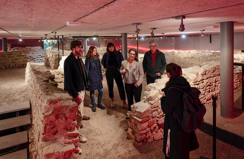 Besucherinnen und Besucher mit Museumsführerin in der versunkenen Offziersküche von Vindonissa. Die Gruppe steht inmitten von Steinmauern.