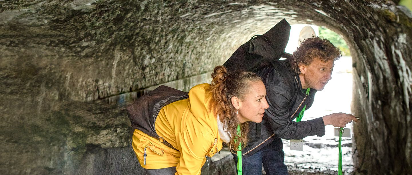 Eine Frau und ein Mann stehen in Freizeitkleidung in einer Höhle auf der Klosterhalbinsel Wettingen (Foxtrail). Sie blicken verwundert an die Wand rechts.