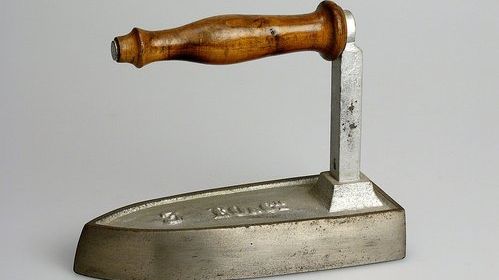 Das Bild zeigt ein Bügeleisen aus Gusseisen mit Holzgriff. Dieses steht vor einem weissen Hintergrund.