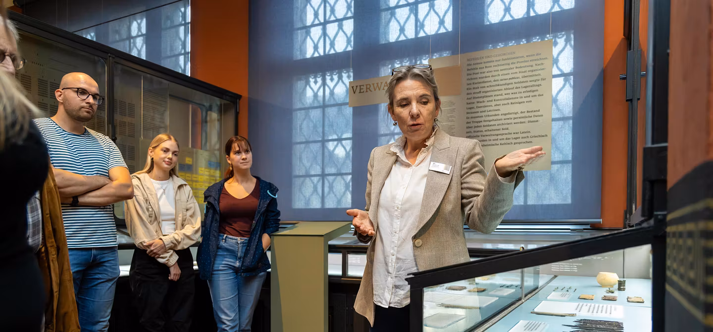 Eine Gruppe Gäste im Vindonissa Museum mit einer Museumsführerin. Diese zeigt auf eine Vitrine