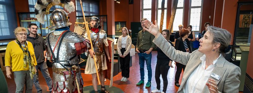 Eine Gruppe Besucherinnen und Besucher steht in der Ausstellung vom Vindonissa Museum (in der Mitte lebensgrosse Legionäre). Eine Museumsführerin steht davor und zeigt auf die beiden Männer.