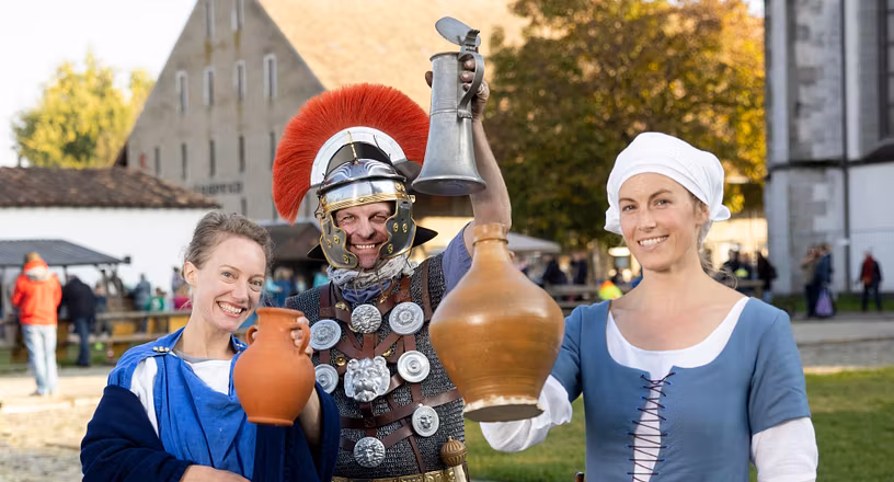Eine Römerin, ein römischer Legionär sowie ein mittelalterliche Magd stehen nebeneinander. Sie blicken mit Weinkrügen in die Kamera und lachten.