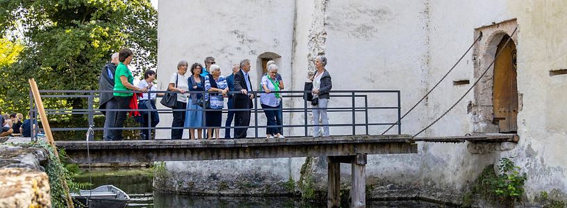Führung auf Schloss Hallwyl: Eine Besuchergruppe steht auf einer Brücke zwischen Hof und dem Eingang zu Schloss Hallwyl, unter der Brücke Wasser des Schlossgrabens und ein Boot. 