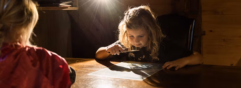 Ein Mädchen untersucht im Kindermuseum von Schloss Lenzburg mit einer Lupe ein Dokument; von hinten erleuchtet ein Scheinwerfer die Szenerie im mystischen Gegenlicht. 
