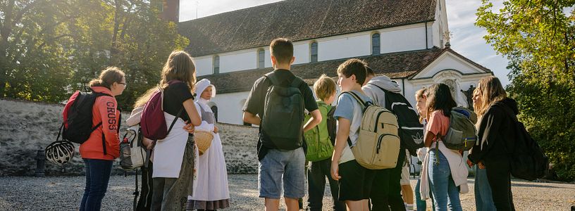 Eine Klostermagd steht mit einer Gruppe Schülerinnen und Schülern vor dem Kloster Wettingen.