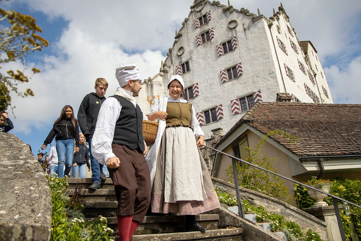 Eine Magd und ein Knecht vor Schloss Wildegg, dahinter eine Schulklasse.