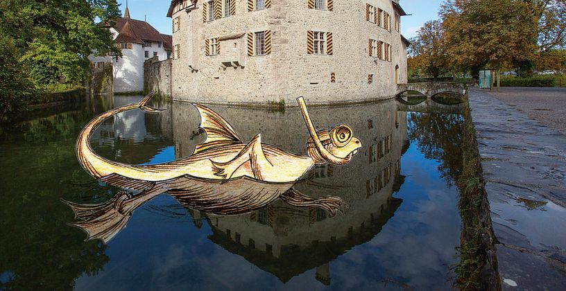 Aussenaufnahme von Schloss Hallwyl mit Aabach im Vordergrund. Im Wasser schwimmt ein Drachentier mit Schnorchel (Illustration), am Himmel ist ein Luftschiff zu sehen (Illustration)