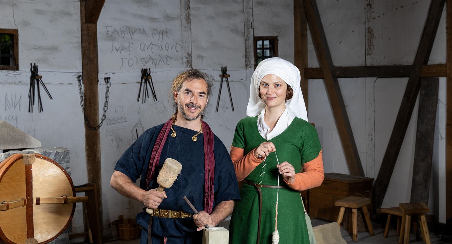 Ein römischer Steinmetz und eine mittelalterlich gekleidete Frau mit Wolle. Beide stehen in einem Werkraum und blicken direkt in die Kamera.