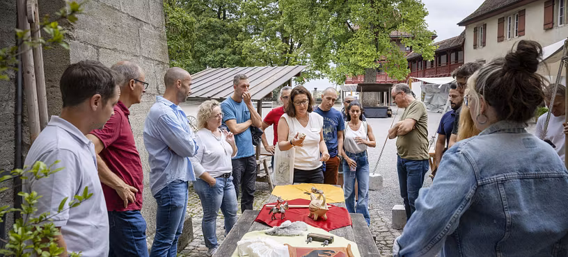 Eine Museumsführerin steht mit Besucherinnen und Besucher um einen grossen Tisch mit verschiedenen Objekten. Im Hintergrund der Hof von Schloss Lenzburg