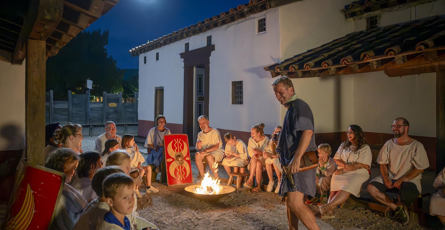 Eine grosse Gruppe mit Erwachsenen und Kindern. Sie sitzen um ein Lagerfeuer im Legionärspfad Vindonissa, in der Mitte brennt ein grosser Feuer. Daneben steht ein römischer Legionär, der lacht. Mystische Stimmung in der Nacht.