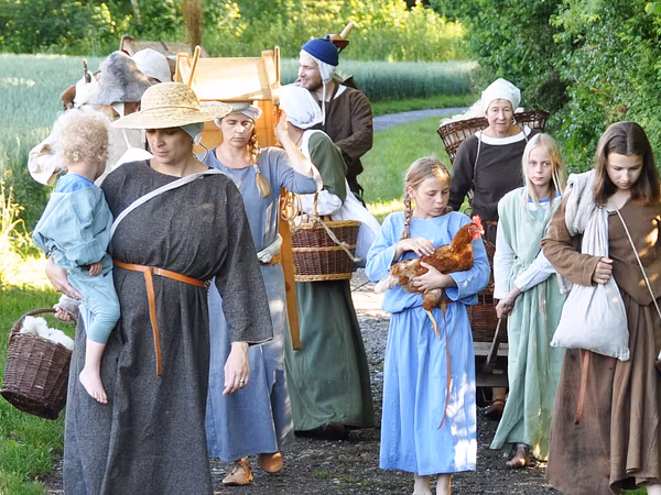 Eine grosse Gruppe Männer, Frauen und Kinder in mittelalterlicher Kleidung. Sie gehen auf einem Weg, ein Kind hält ein Huhn in den Händen.
