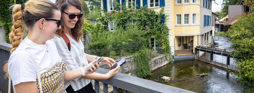 Zwei Frauen stehen mit Handys an einem Bach, im Hintergrund das "Klein Venedig" von Lenzburg
