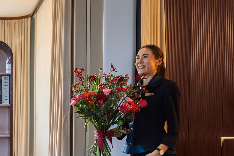 Eine elegant gekleidete Frau schreitet mit einem Blumenstrauss durch den Aufenthaltsraum eines Hotels.