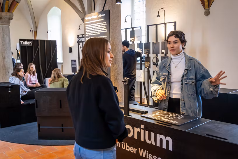 Ausstellung "Parlatorium auf der Klosterhalbinsel Wettingen": Zwei Schülerinnen diskutieren in einem grossen Raum; dahinter weitere Personen sitzend.