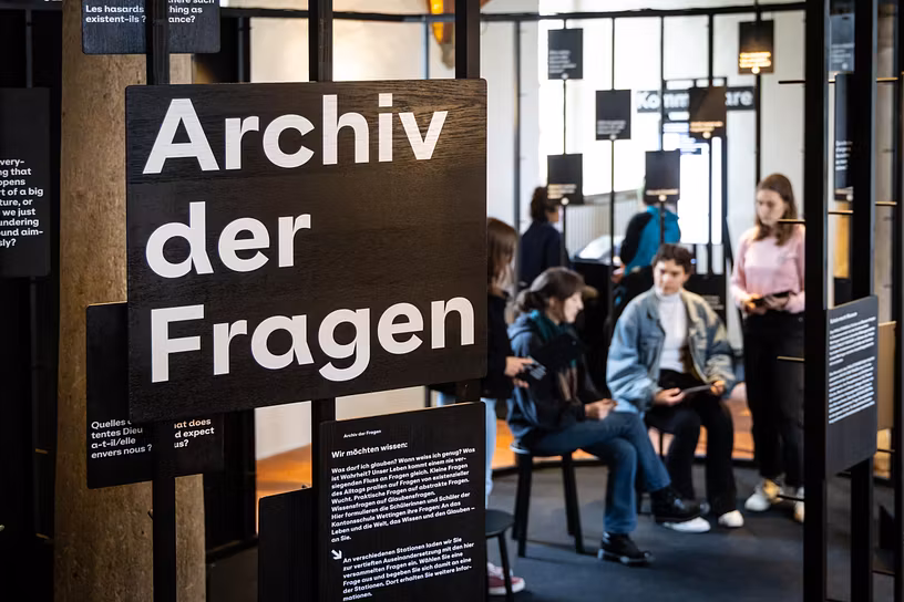 Ausstellungsraum im Parlatorium der Klosterhalbinsel Wettingen: Ein grosses Schild mit dem Text "Archiv der Fragen", im Hintergrund sind mehrere Personen zu sehen, die diskutieren.