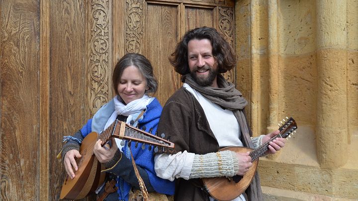 Ein Mann und eine Frau stehen mit je einem Musikinstrument vor einer Tür. Beide sind mittelalterlich gekleidet.