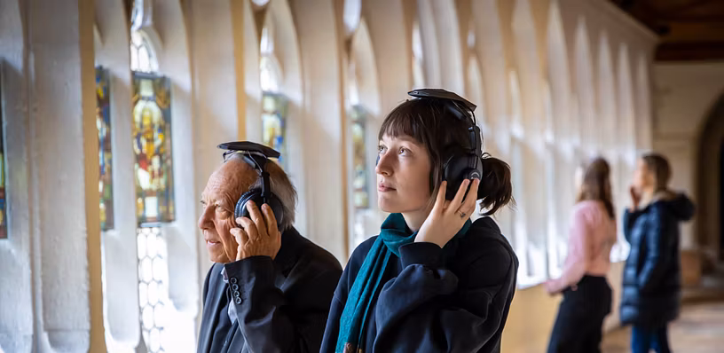 Ein Mann und eine Frau stehen im Kreuzgang der Klosterhalbinsel Wettingen. Beide tragen grosse Kopfhörer. Im Hintergrund weitere Personen.
