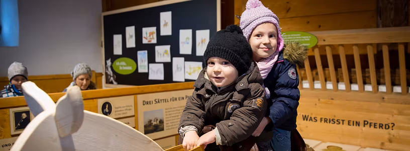 Mädchen und Junge reiten auf einem Holzpferd im Pferdestall auf Schloss Wildegg