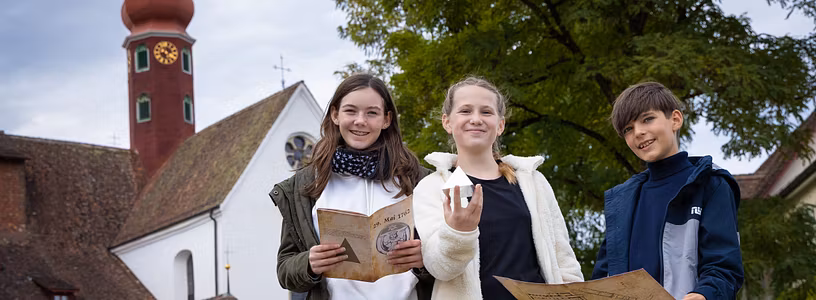 Zwei Mädchen und ein Bub stehen gemeinsam vor dem Kloster Wettingen und blicken direkt in die Kamera. Sie halten eine Landkarte, Broschüre sowie eine Pyramide in den Händen.