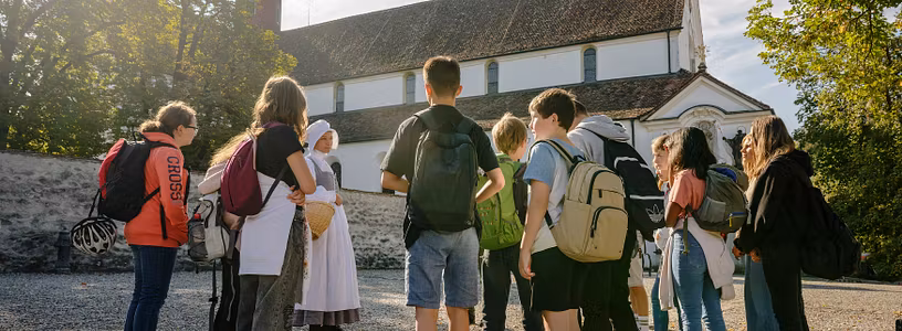 Eine Klostermagd steht mit einer Gruppe Schülerinnen und Schülern vor dem Kloster Wettingen.