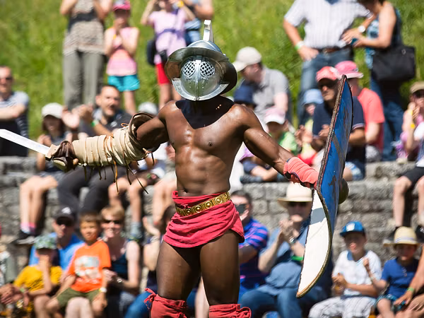 Ein Gladiator mit Schwert, Schild und Helm im Amphitheater vom Legionärspfad Vindonissa. Im Hintergrund ist unscharf das Publikum zu erkennen.