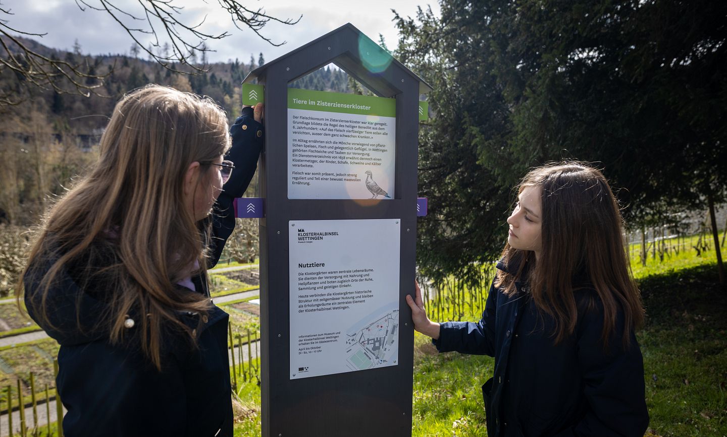 Zwei Mädchen stehen vor einer Info-Stele mit Textinformationen inmitten dem Garten der Klosterhalbinsel Wettingen.