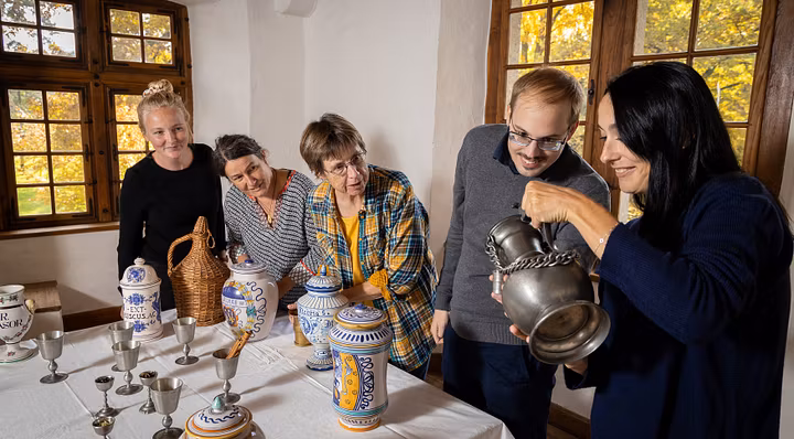 Eine Besuchergruppe in einem historischen Raum von Schloss Hallwyl. Die Gruppe steht um einen Tisch herum, auf dem Gefässe stehen. Eine Frau geisst eine Flüssigkeit aus einem Metallkrug in einen Metallbecher.