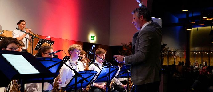 Im Bild sind links mehrere musizierende Personen zu sehen. Alle spielen verschiedene Blasinstrumente und haben ein weisses Hemd an. Vor den sitzenden Bandmitglieder steht der Dirigent in einem grauen Jackett.  