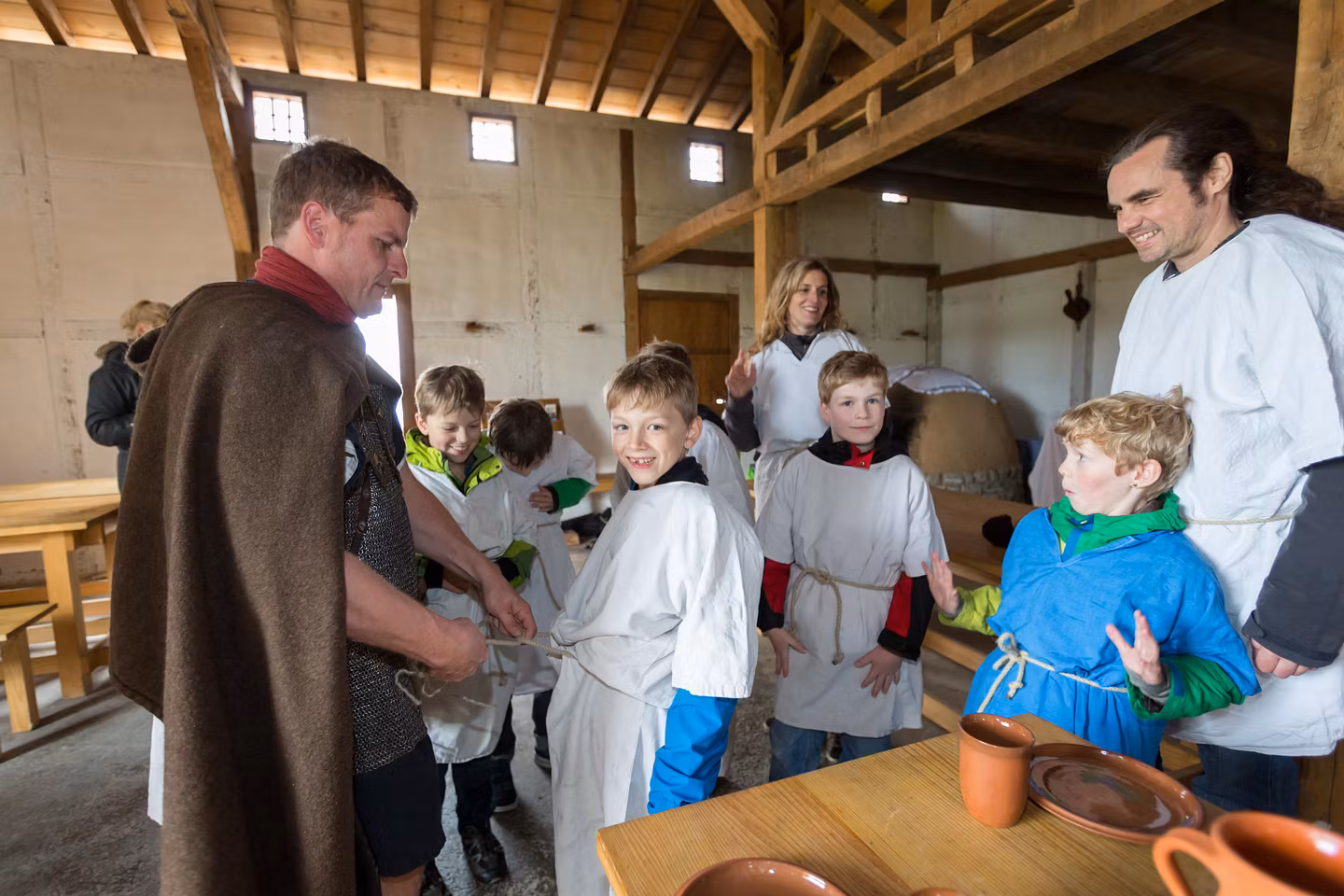 Ein römisch gekleideter Mann mit mehreren Schülerinnen und Schülern. Der Mann schnürt bei einem Schüler eine römische Tunika zu.