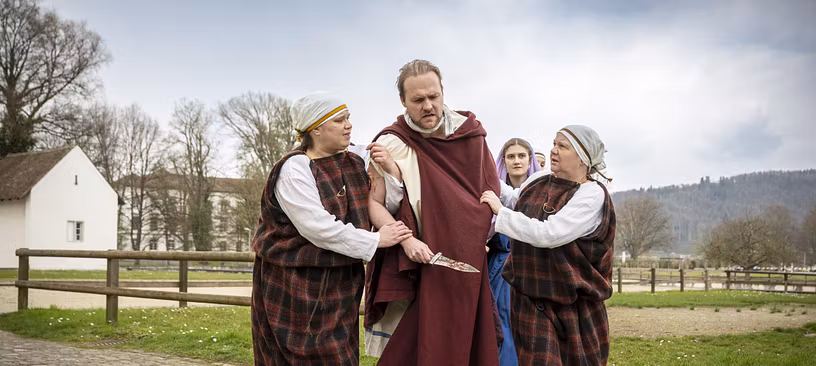 Mehrere Schauspieler in historischer Kleidung. Sie stehen nahe zusammen, der Mann in der Mitte hält einen Dolch in der Hand.