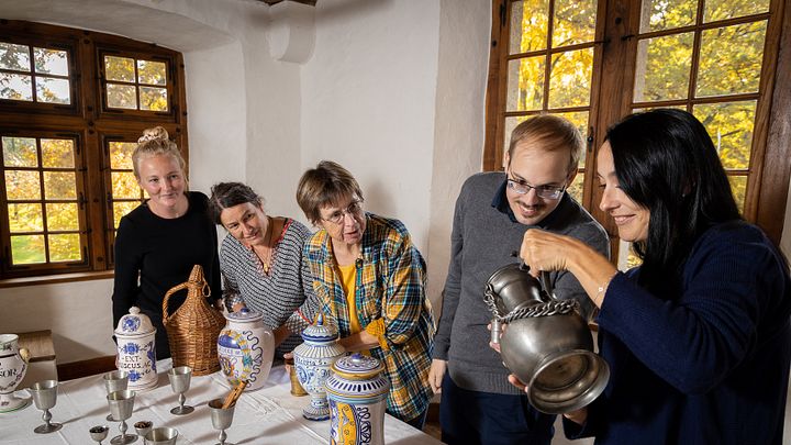 Eine Besuchergruppe in einem historischen Raum von Schloss Hallwyl. Die Gruppe steht um einen Tisch herum, auf dem Gefässe stehen. Eine Frau geisst eine Flüssigkeit aus einem Metallkrug in einen Metallbecher.