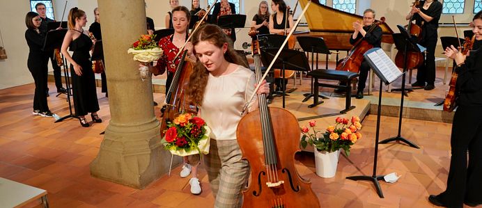 Viele Personen mit Streichinstrumenten am Stehen nach einem Konzert. Zwei Personen laufen mit Blume und Instrument in der Hand davon.