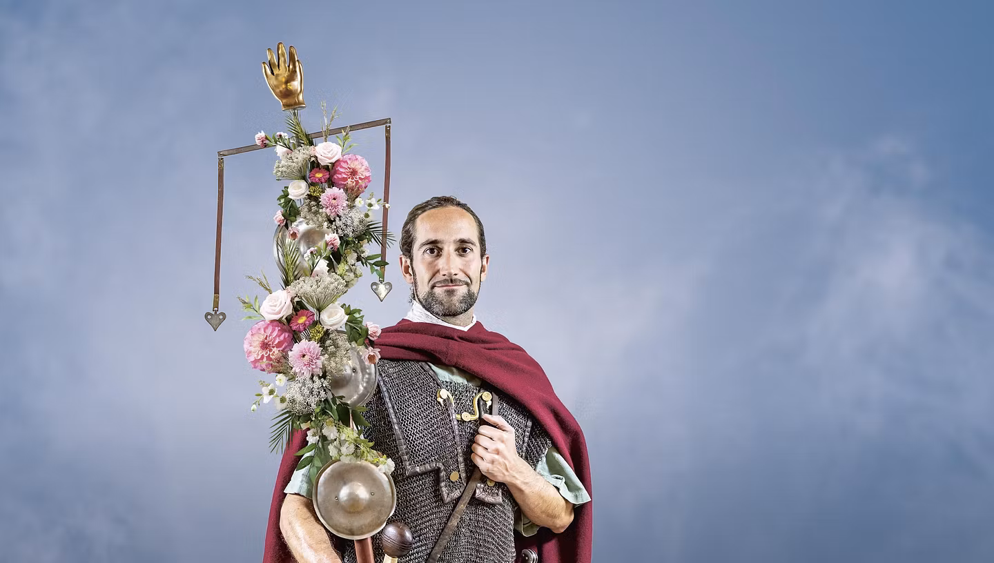 Ein römischer Legionär vor blauem Hintergrund. Er trägt eine mit Blumen geschmückte Standarte in der rechten Hand. 