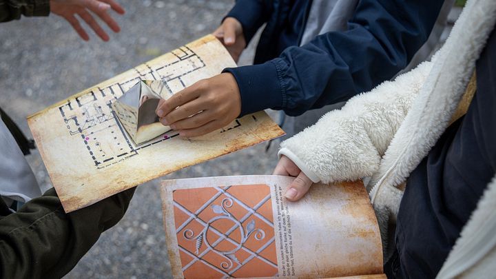Detailaufnahmen von einem mysteriösen Plan und einem Buch, die auf den Knien von zwei Schülern liegen.